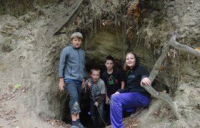 Vier Kinder stehen am Eingang einer kleinen H&ouml;hle im Wald.