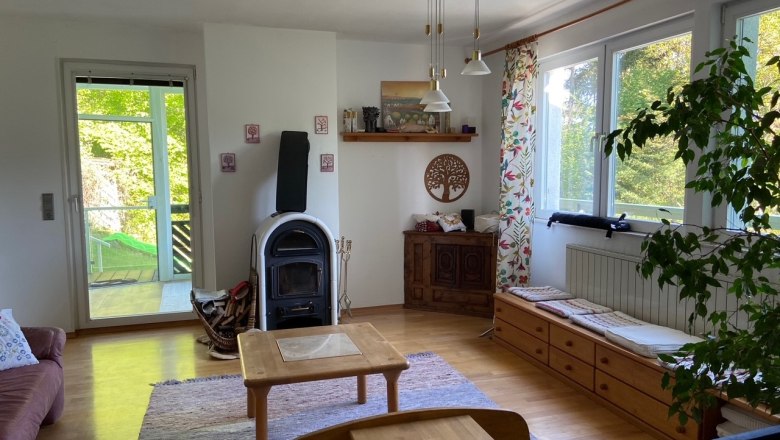 Ein gem&uuml;tliches Wohnzimmer mit Holzofen, Holzm&ouml;beln und gro&szlig;en Fenstern mit Blick ins Gr&uuml;ne.