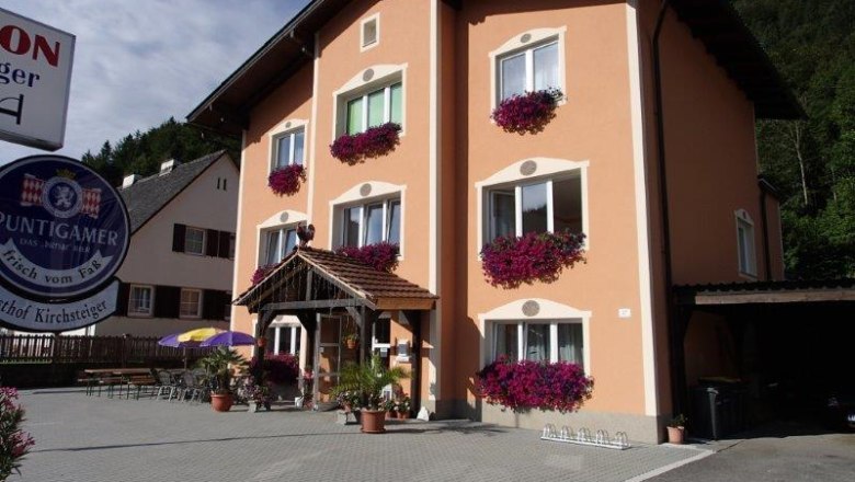 Pension-Gasthof Kirchsteiger, © zVg Ein dreistöckiges Gebäude mit orangefarbener Fassade und Blumenkästen vor den Fenstern. Ein Schild mit der Aufschrift 'Puntigamer' ist sichtbar.
