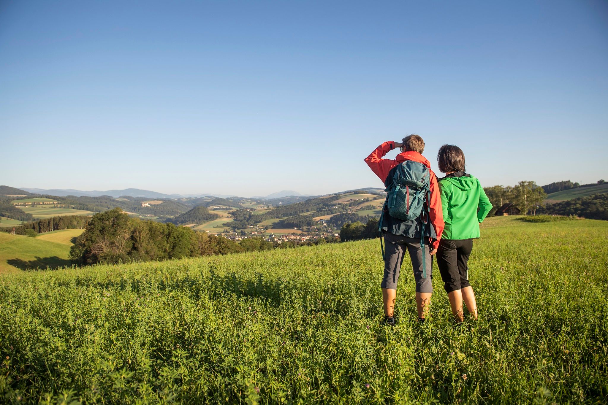 Zwei Personen stehen auf einer Wiese und blicken in die Ferne über eine hügelige Landschaft.
