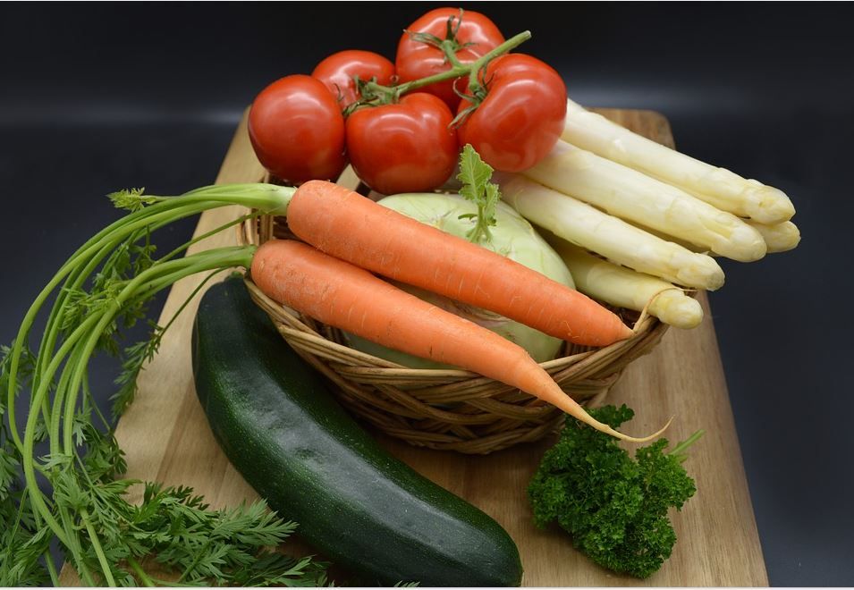 Korb mit frischem Gemüse auf einem Holzbrett: Karotten, Tomaten, weißer Spargel, Zucchi ni, Kohl und Kräutervor dunklem Hintergrund.