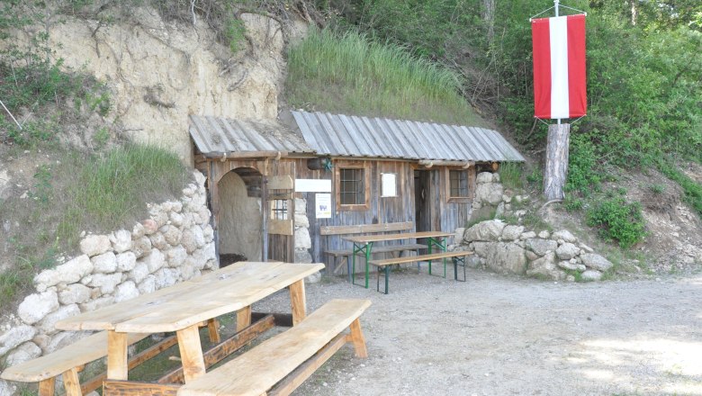 Eine rustikale Holzh&uuml;tte mit Tisch und B&auml;nken davor, eingebaut in einen H&uuml;gel. Eine rot-wei&szlig;e Fahne h&auml;ngt an einem Baum.