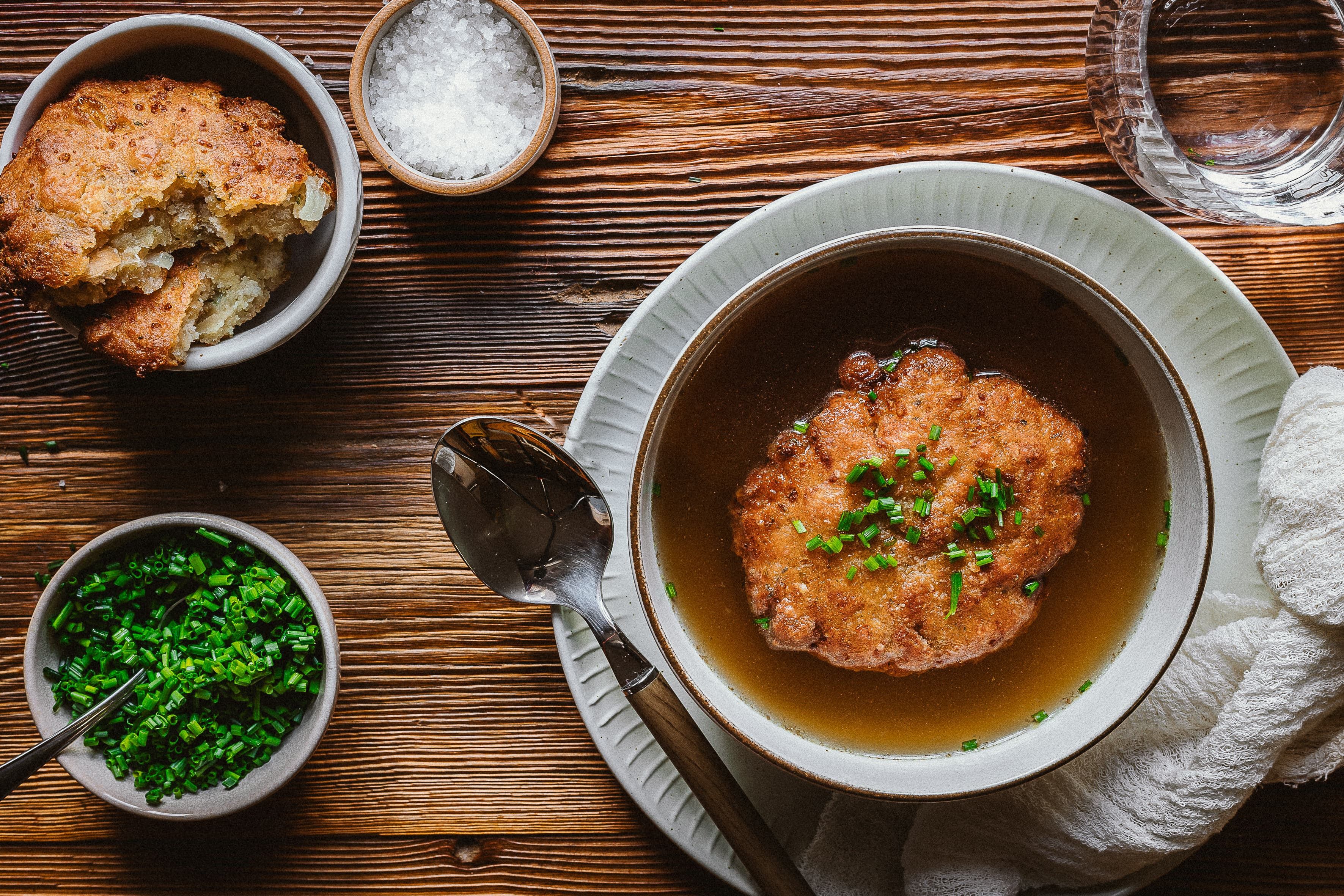 Kaspressknödelsuppe in einer Schüssel auf einem Holztisch, mit Schnittlauch und Salz in kleinen Schalen daneben.