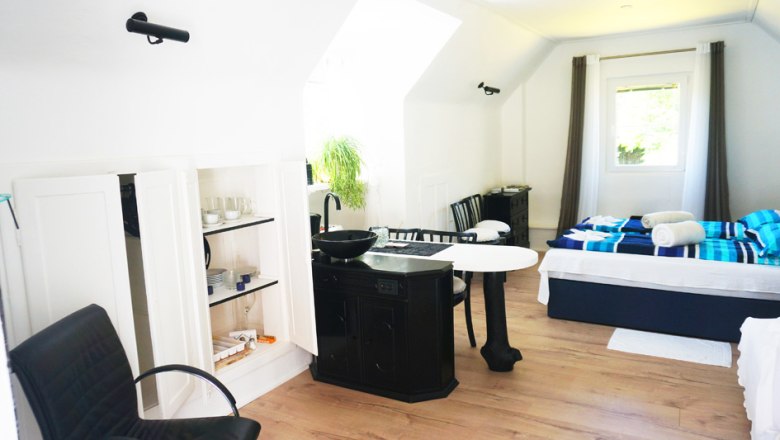 Innenansicht eines hellen Ferienhauses mit Bett, Tisch, Stühlen und Küchenzeile.