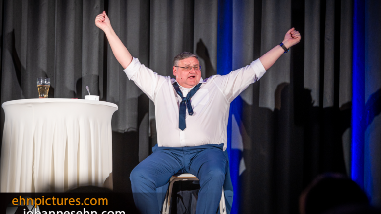 Ein Mann sitzt auf einer B&uuml;hne mit erhobenen Armen, neben ihm ein Tisch mit einem Glas.