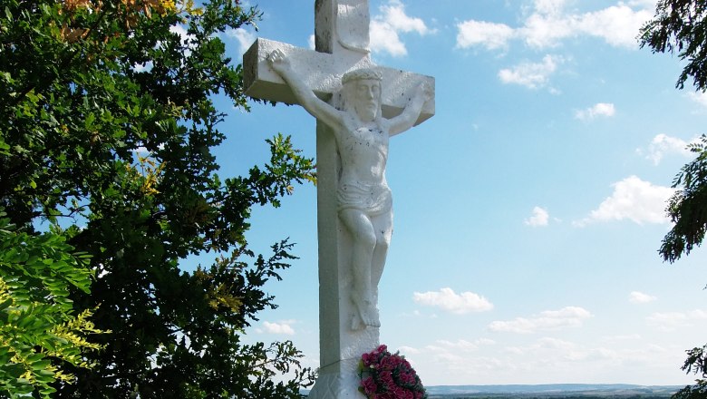 Wei&szlig;es Steinkreuz mit Jesusfigur und Blumenkranz, umgeben von B&auml;umen und Landschaft im Hintergrund.