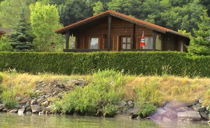 Ein Holzferienhaus mit rotem Dach, umgeben von B&auml;umen und Str&auml;uchern, steht am Ufer eines Gew&auml;ssers.