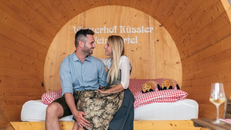 Ein Paar sitzt l&auml;chelnd in einem gro&szlig;en Holzfass mit der Aufschrift 'Winzerhof K&uuml;ssler Weinviertel'.