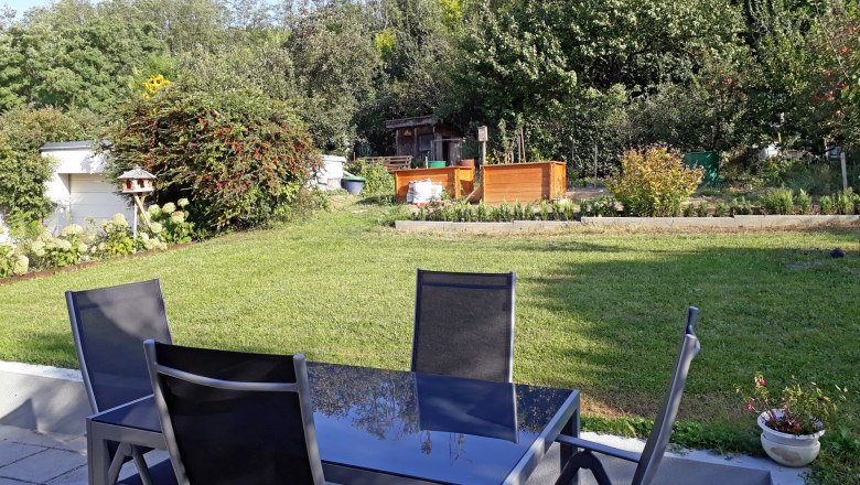 Terrasse mit Tisch und St&uuml;hlen, Blick auf einen Garten mit Rasen, B&auml;umen und Hochbeeten.