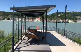 Anlegestelle mit Sitzbänken am Fluss, im Hintergrund eine Stadt und eine Basilika auf einem Hügel.