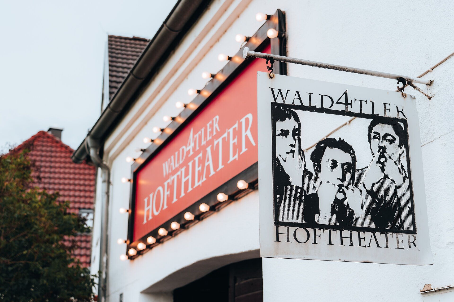 Außenansicht des Wald4tler Hoftheaters mit beleuchtetem Schild und einem Bild von drei Personen.