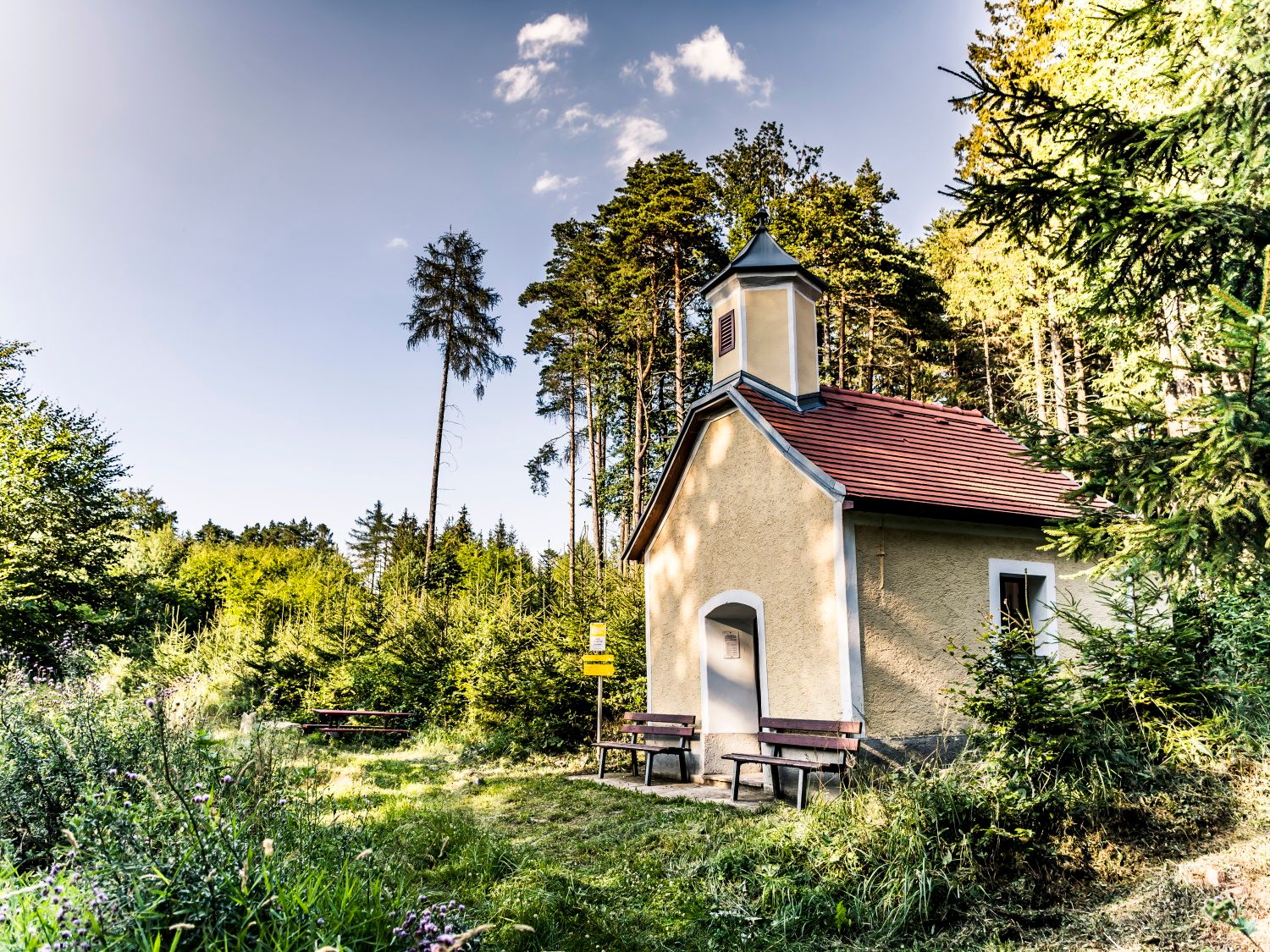 Kleine Kapelle im Wald mit Bänken davor, umgeben von Bäumen und Wiese.
