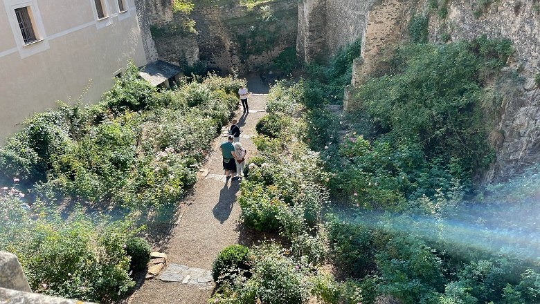 Ein Garten mit Wegen und Pflanzen in einer alten Burganlage, Menschen spazieren.
