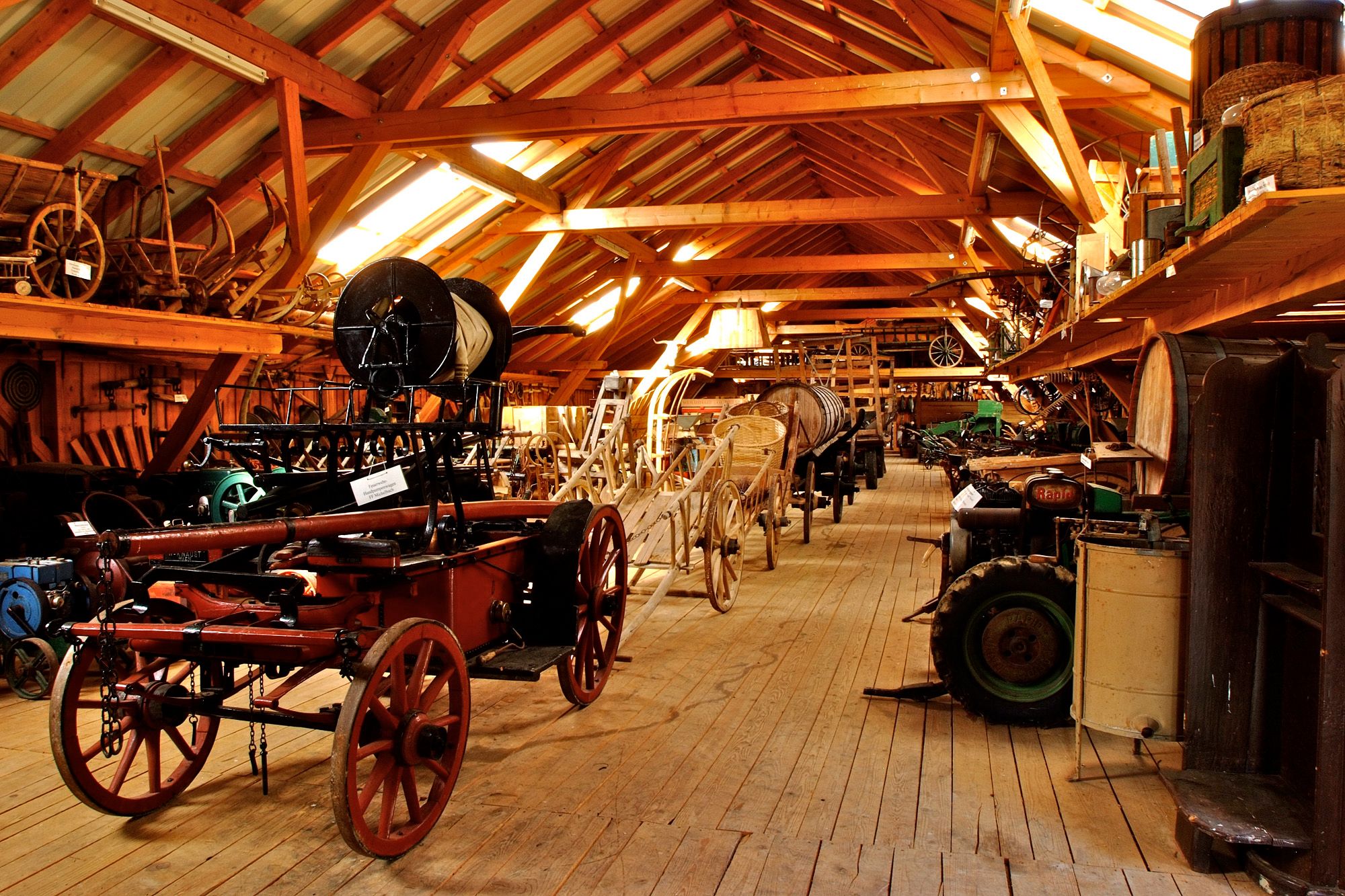 Innenansicht eines Museums mit alten landwirtschaftlichen Geräten und Wagen auf Holzboden.