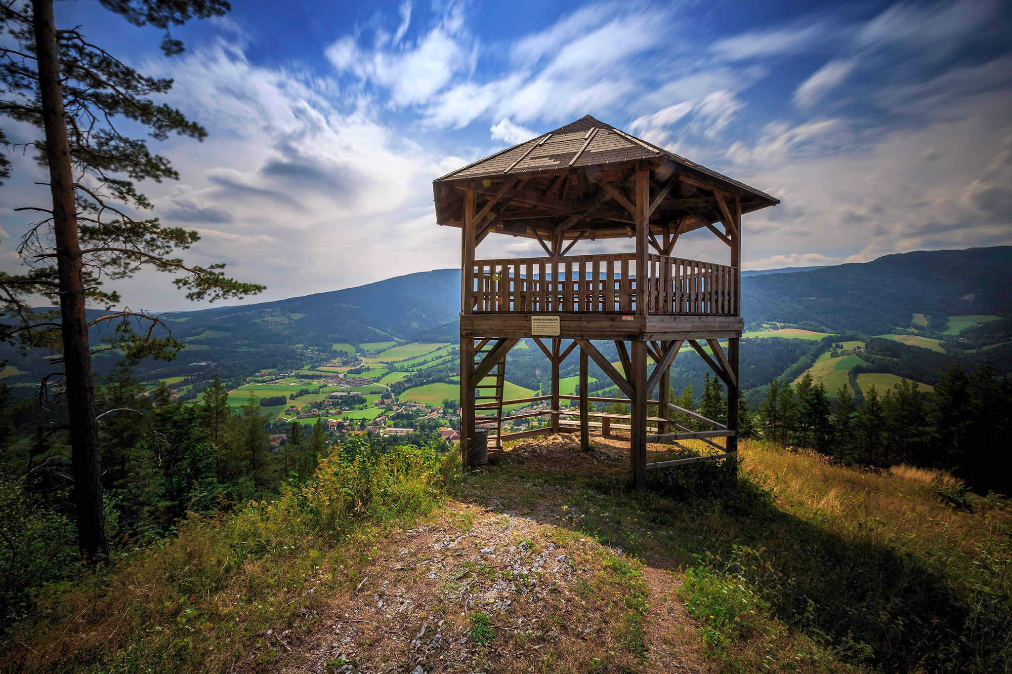 Holzaussichtsturm mit Blick auf grüne Täler und Berge unter blauem Himmel.