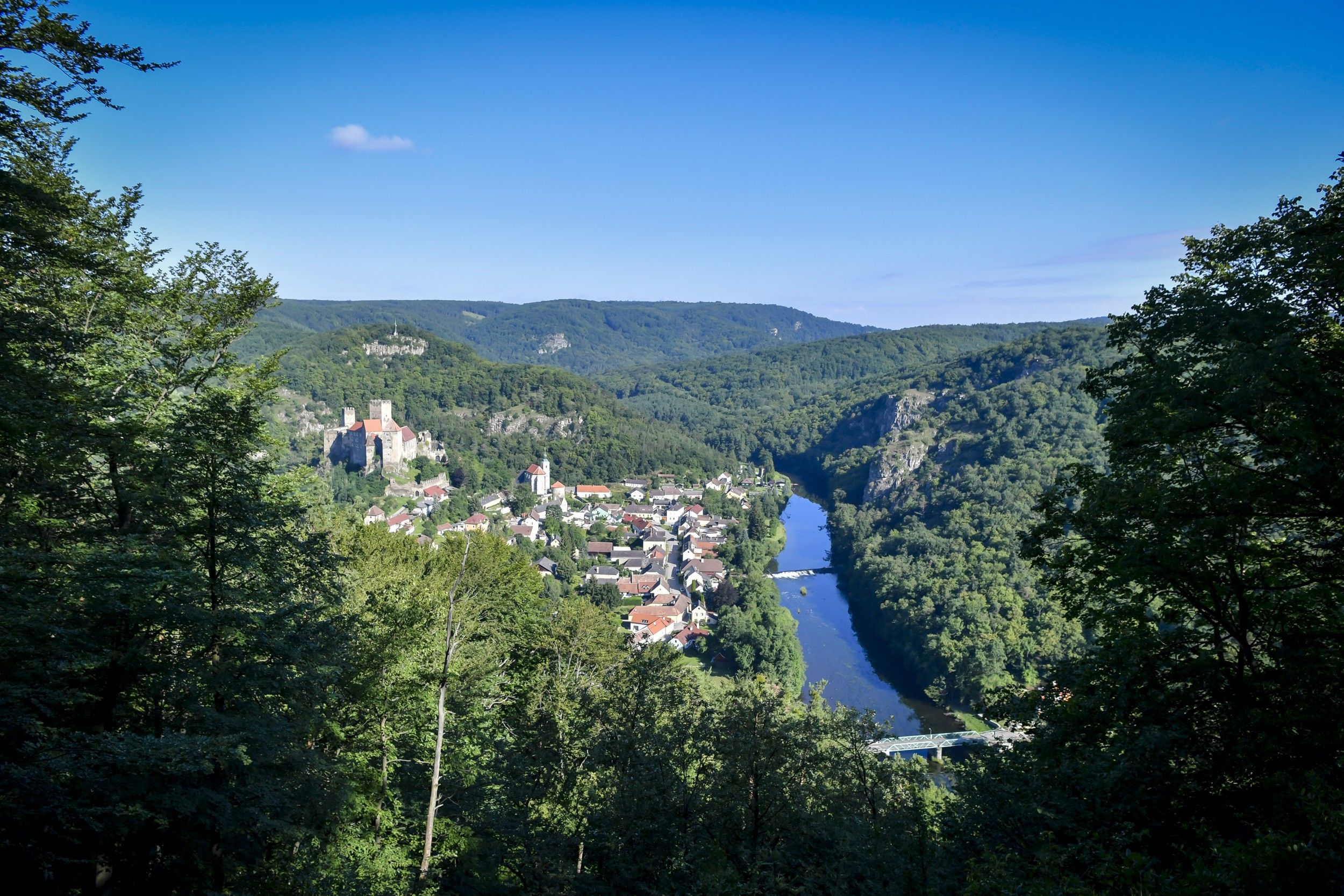 Panoramablick auf eine Burg und ein Dorf in einem bewaldeten Tal mit Fluss.