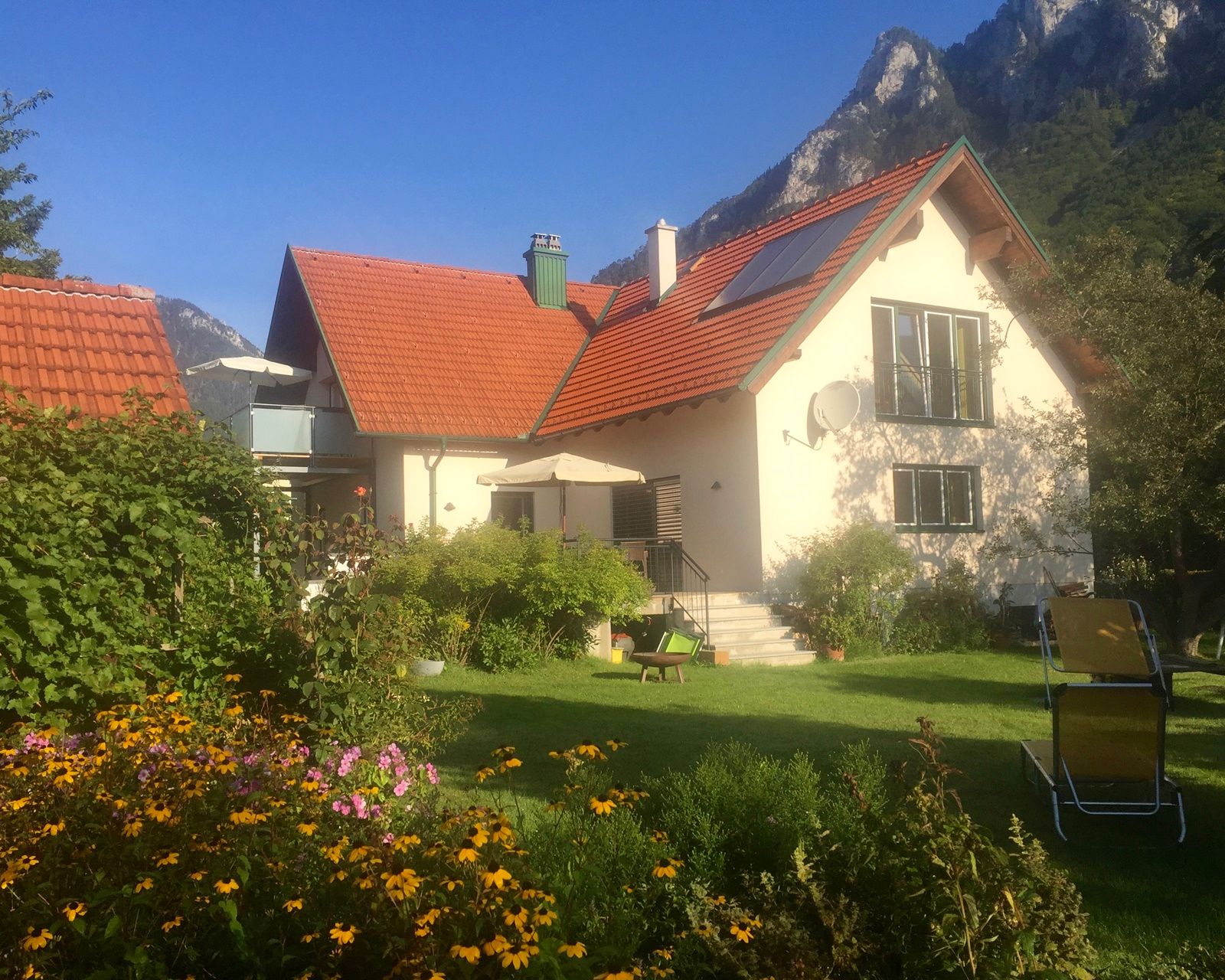 Ein Haus mit rotem Dach, umgeben von einem Garten mit Blumen und Liegestühlen, vor einer Bergkulisse.