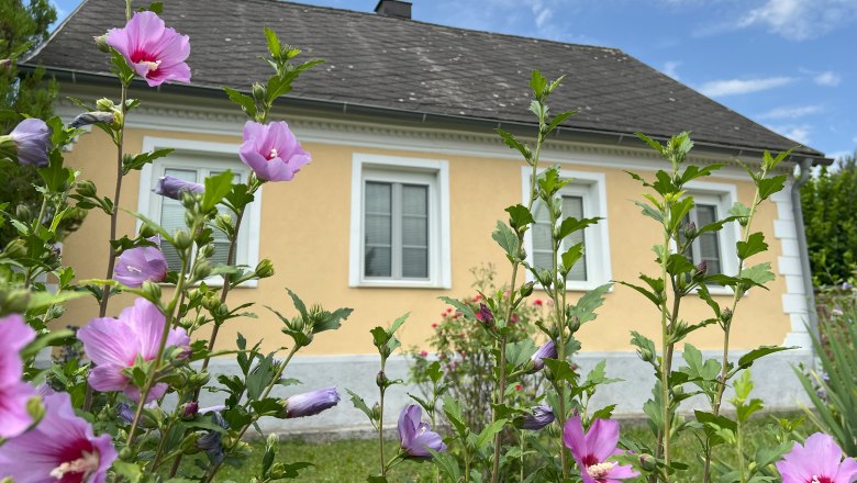 Herzlich Willkommen, © Waltraud Forstner Ein gelbes Haus mit weißen Fensterrahmen, umgeben von blühenden rosa Blumen im Vordergrund.