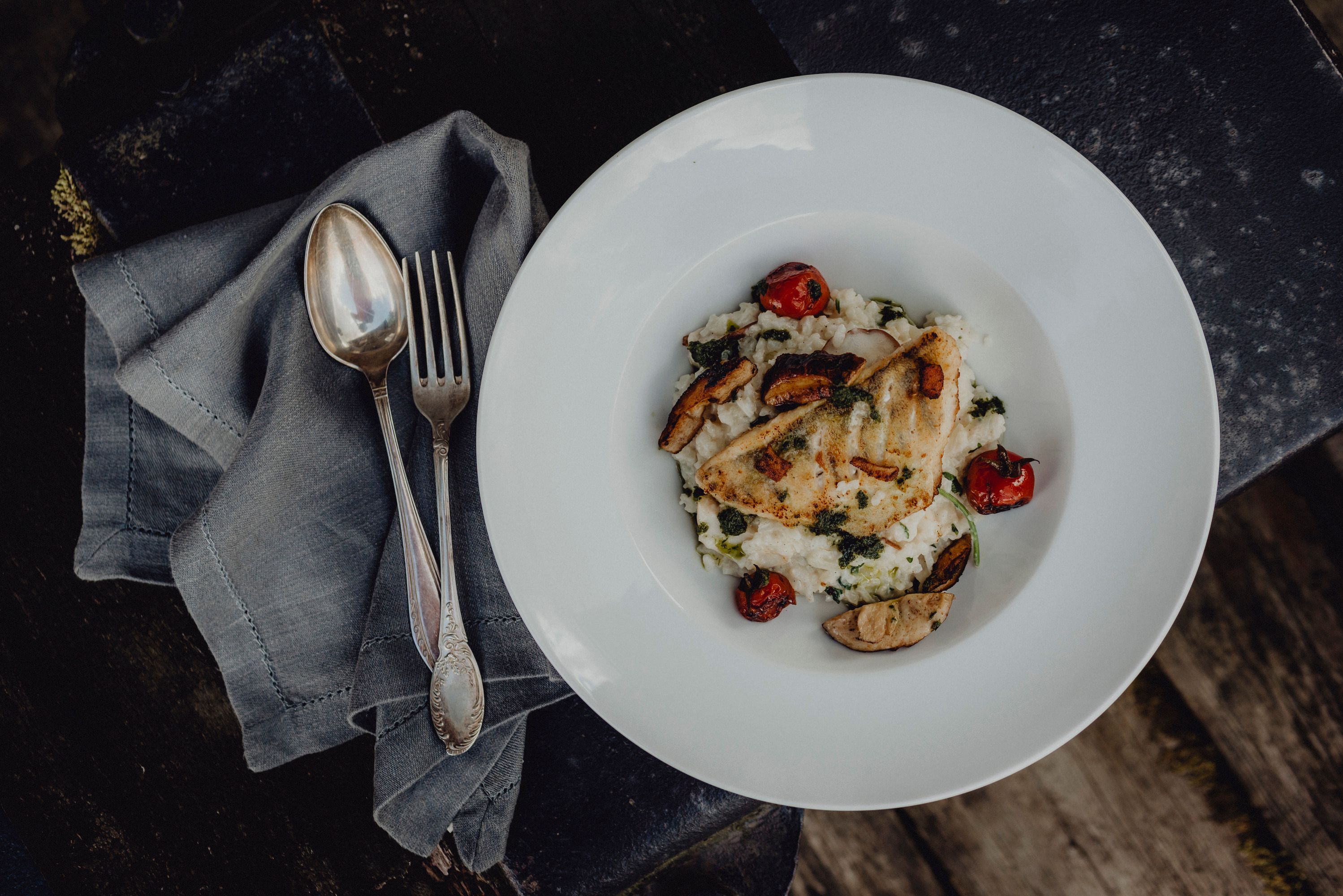 Ein Teller mit Fischgericht, serviert auf Risotto mit Tomaten und Pilzen, daneben Besteck auf einer Serviette.
