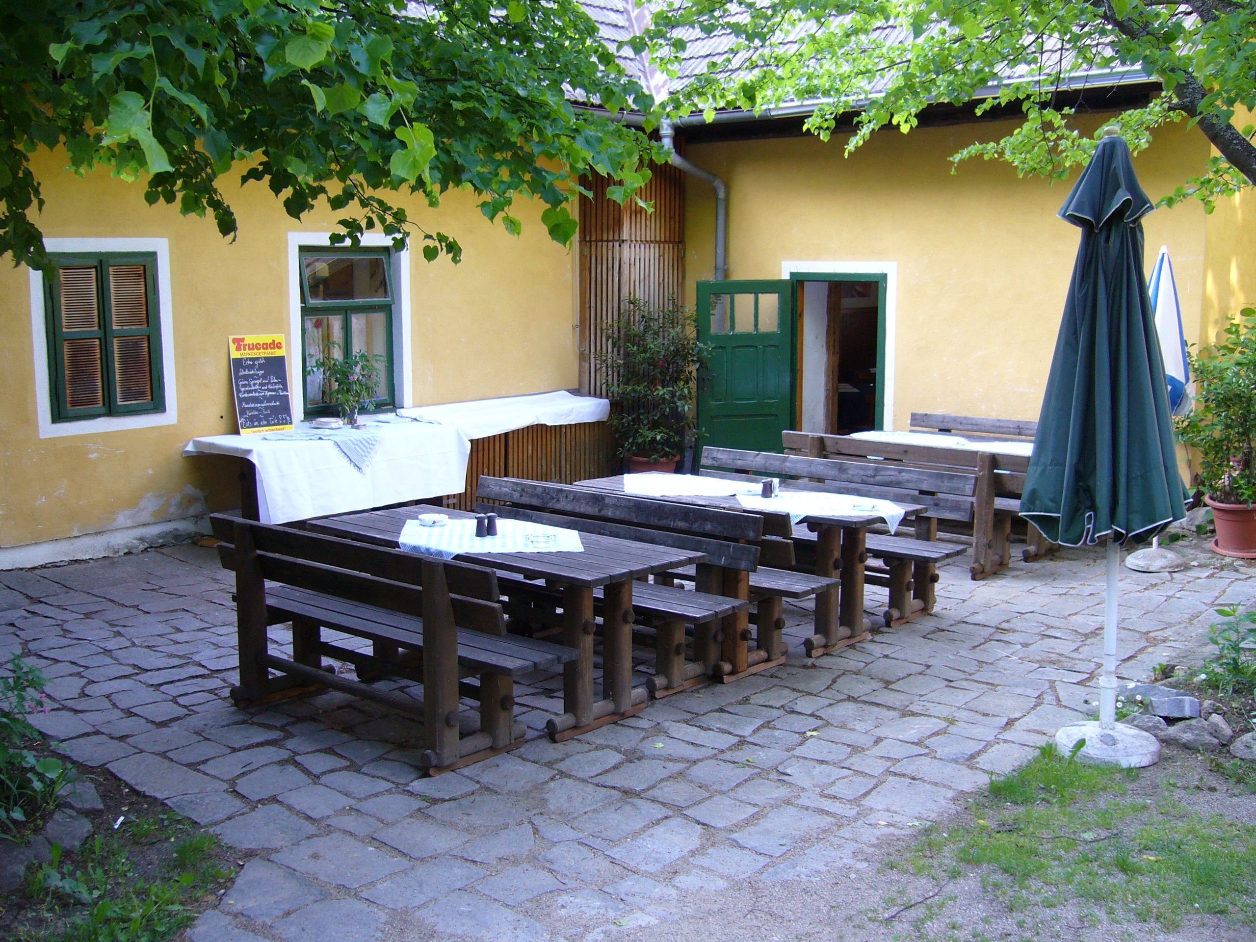 Innenhof eines Gasthauses mit Holzbänken und Tischen, gelbe Wand, grüner Tür und Fenster, Sonnenschirm und Pflanzen.