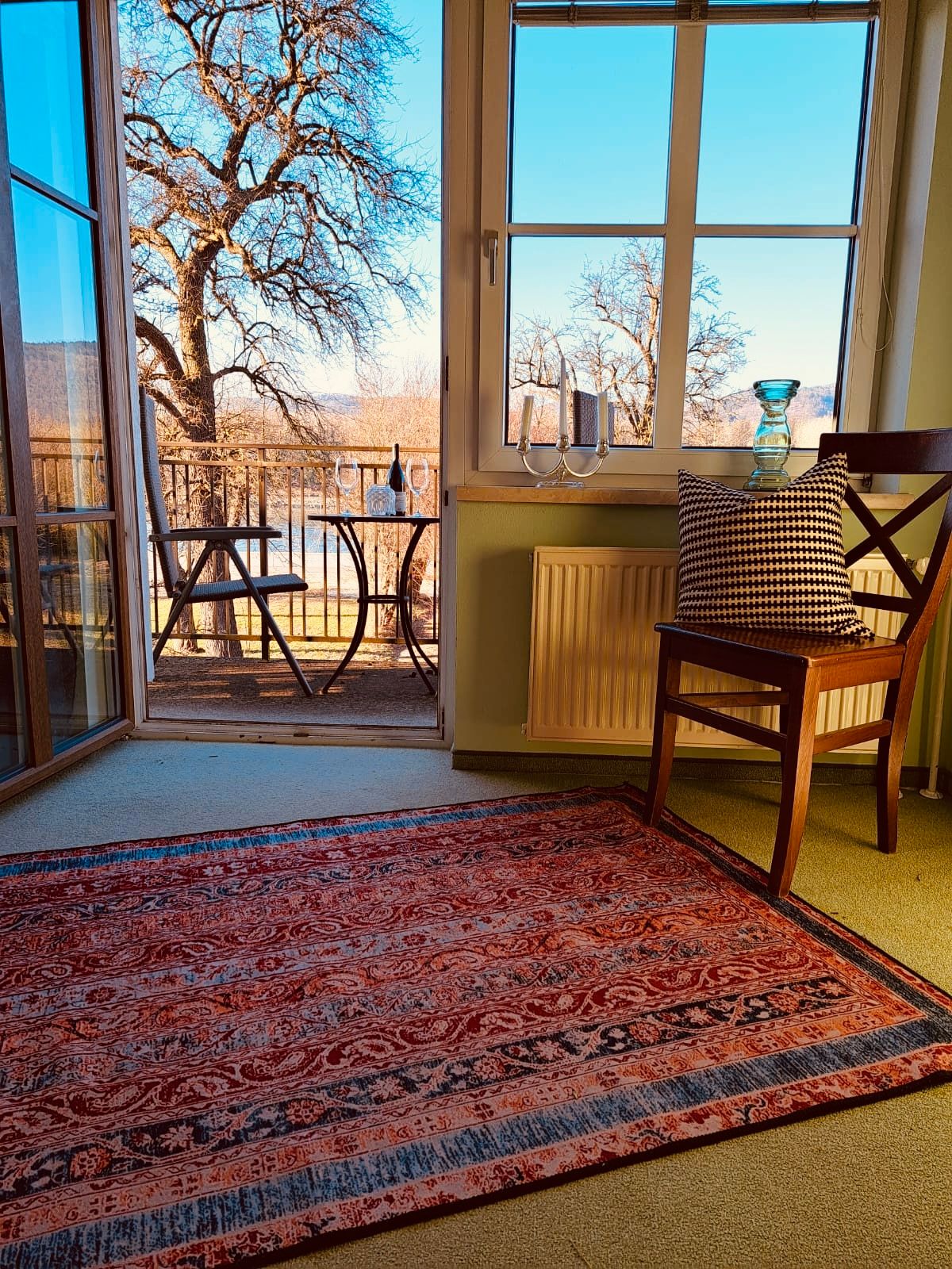 Ein Zimmer mit Teppich, Stuhl und Blick auf einen Balkon mit Tisch und Stühlen, Bäume im Hintergrund.