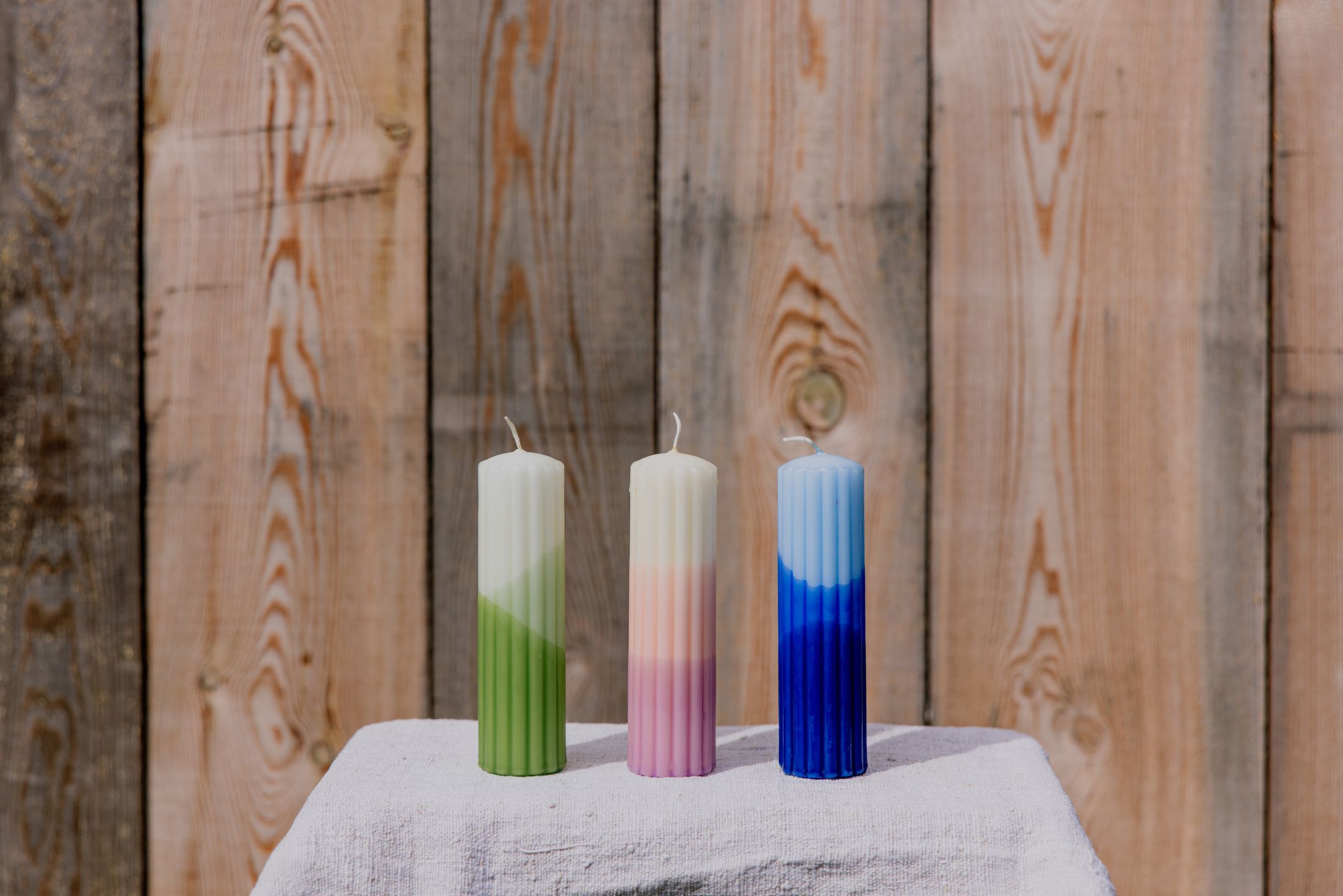 Drei bunte Kerzen auf einem Tisch vor einer Holzwand.