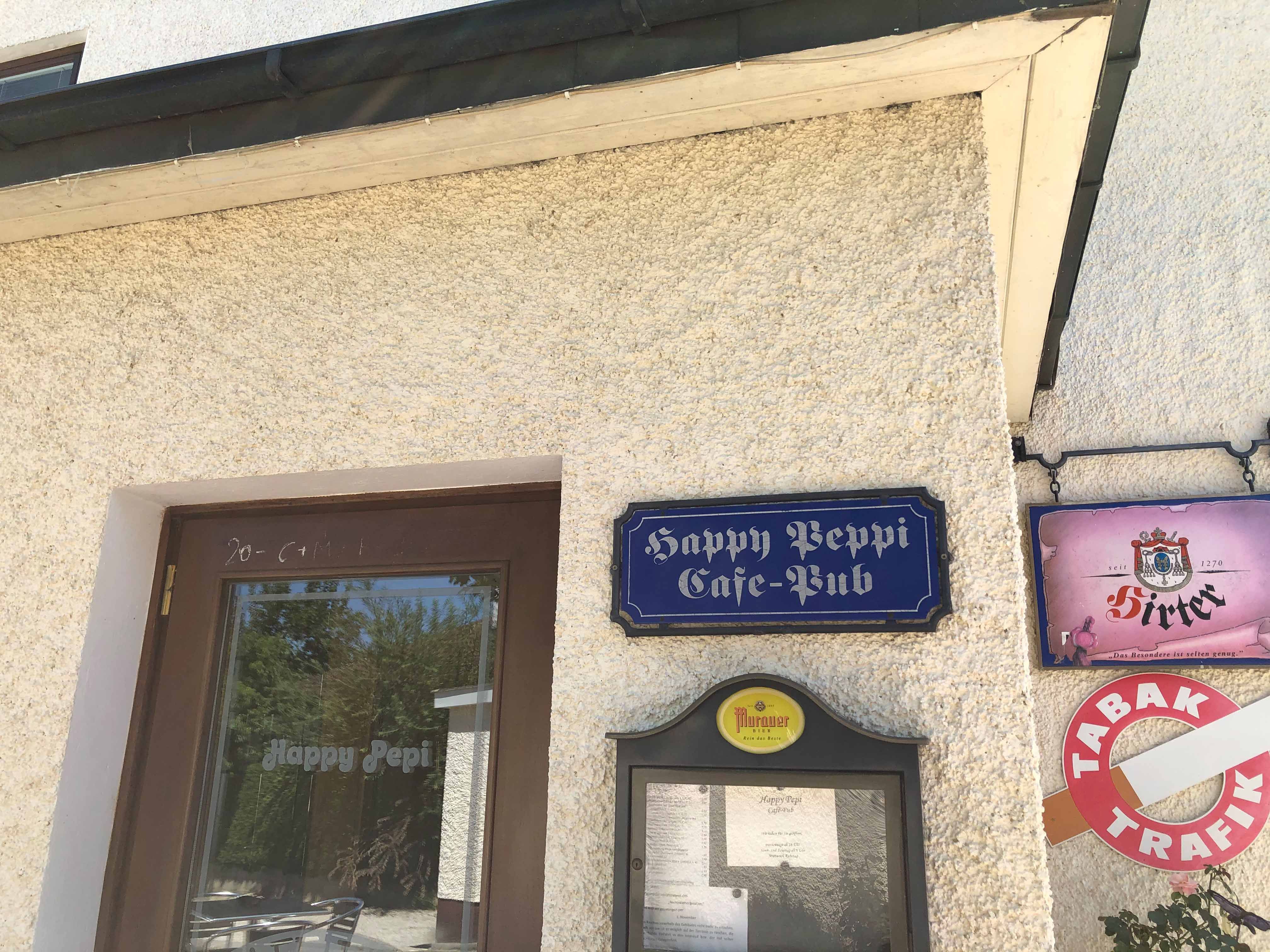 Eingang des Cafe-Pubs Happy Pepi mit Schildern und Menü an der Wand.