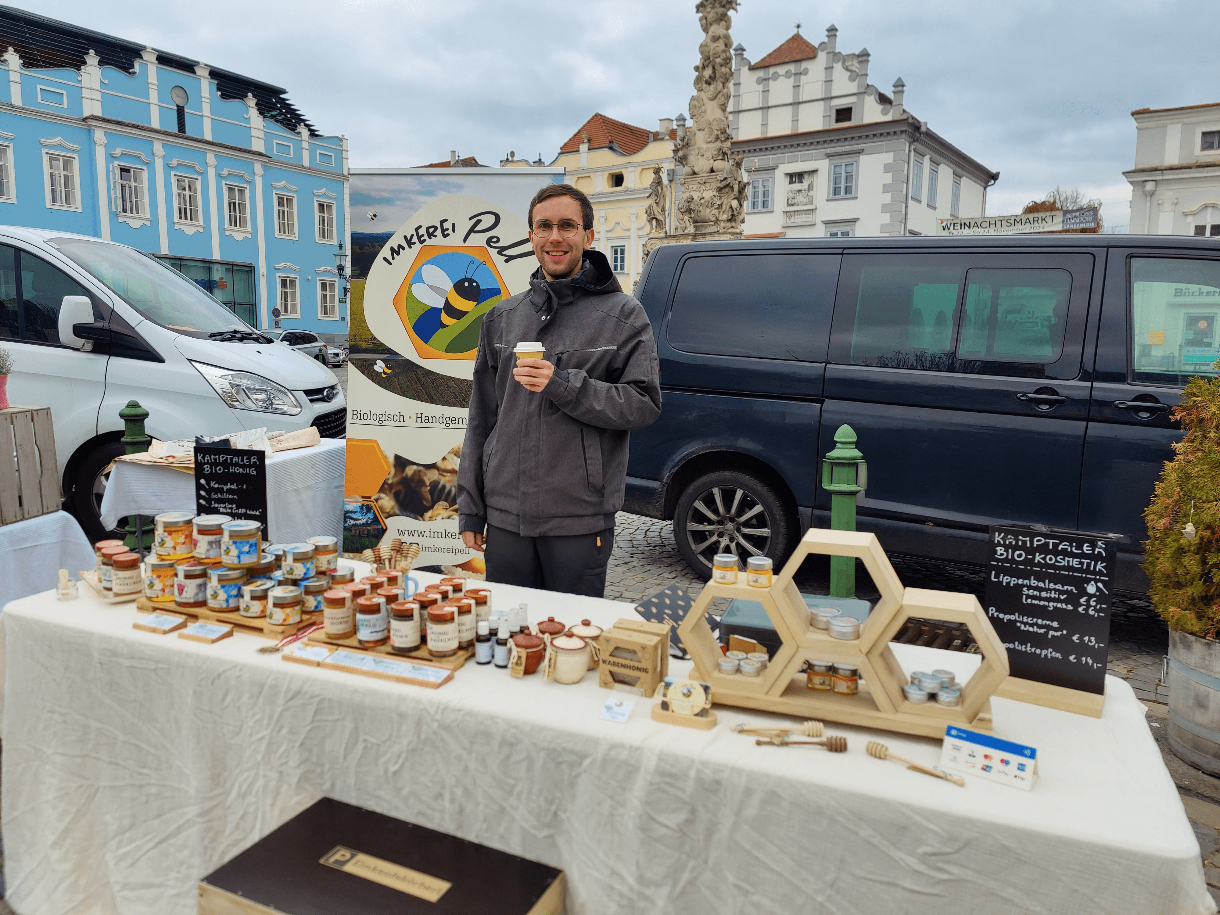 Ein Mann steht an einem Marktstand mit Honigprodukten und hält einen Becher. Im Hintergrund sind ein Van und Gebäude zu sehen.