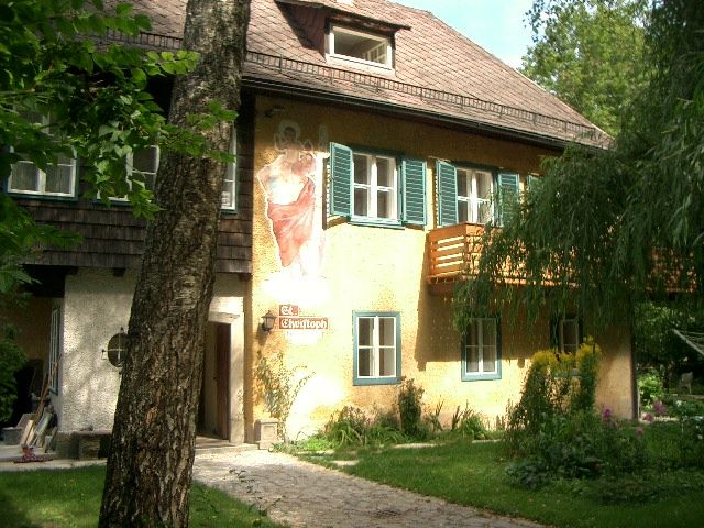 Ein zweistöckiges Haus mit gelber Fassade, grünen Fensterläden und einem Wandgemälde. Ein Baum steht im Vordergrund.