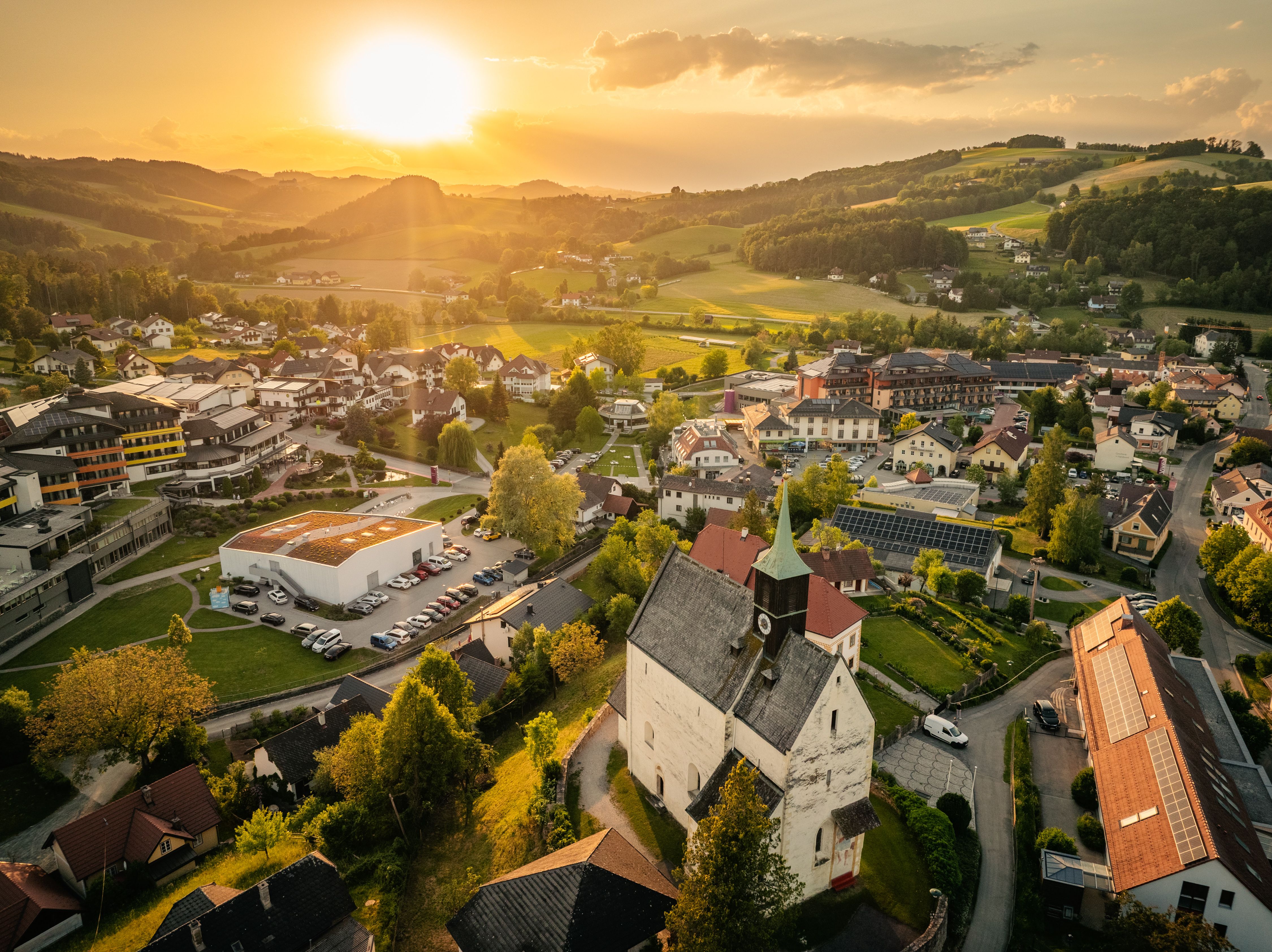 Luftaufnahme von Bad Schönau bei Sonnenuntergang mit Wehrkirche im Vordergrund und Hügeln im Hintergrund.