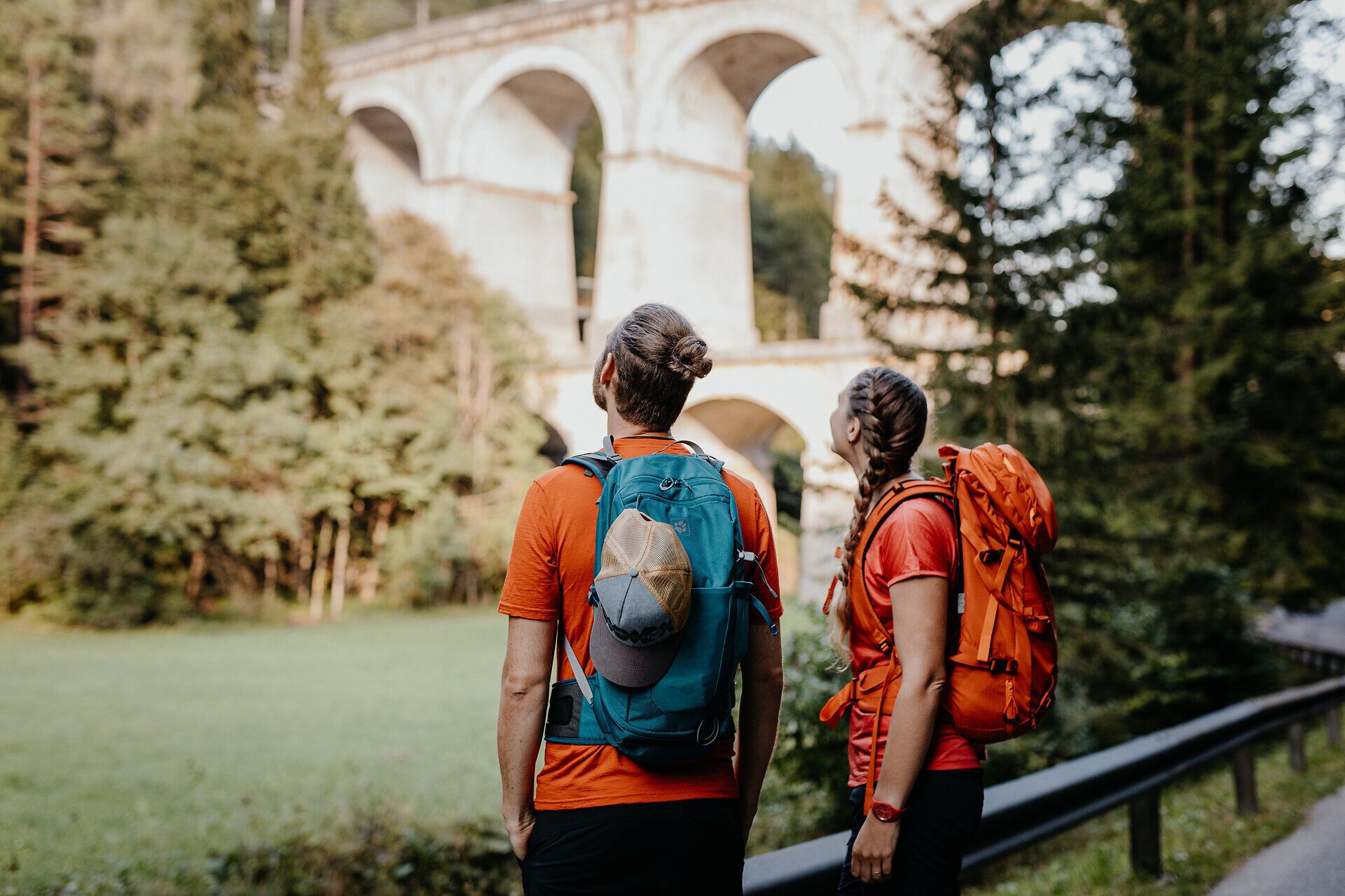 Zwei Wandernde mit Rucksäcken stehen am Ufer eines Flusses und blicken hinauf zum steinernen Viadukt der Semmeringbahn.