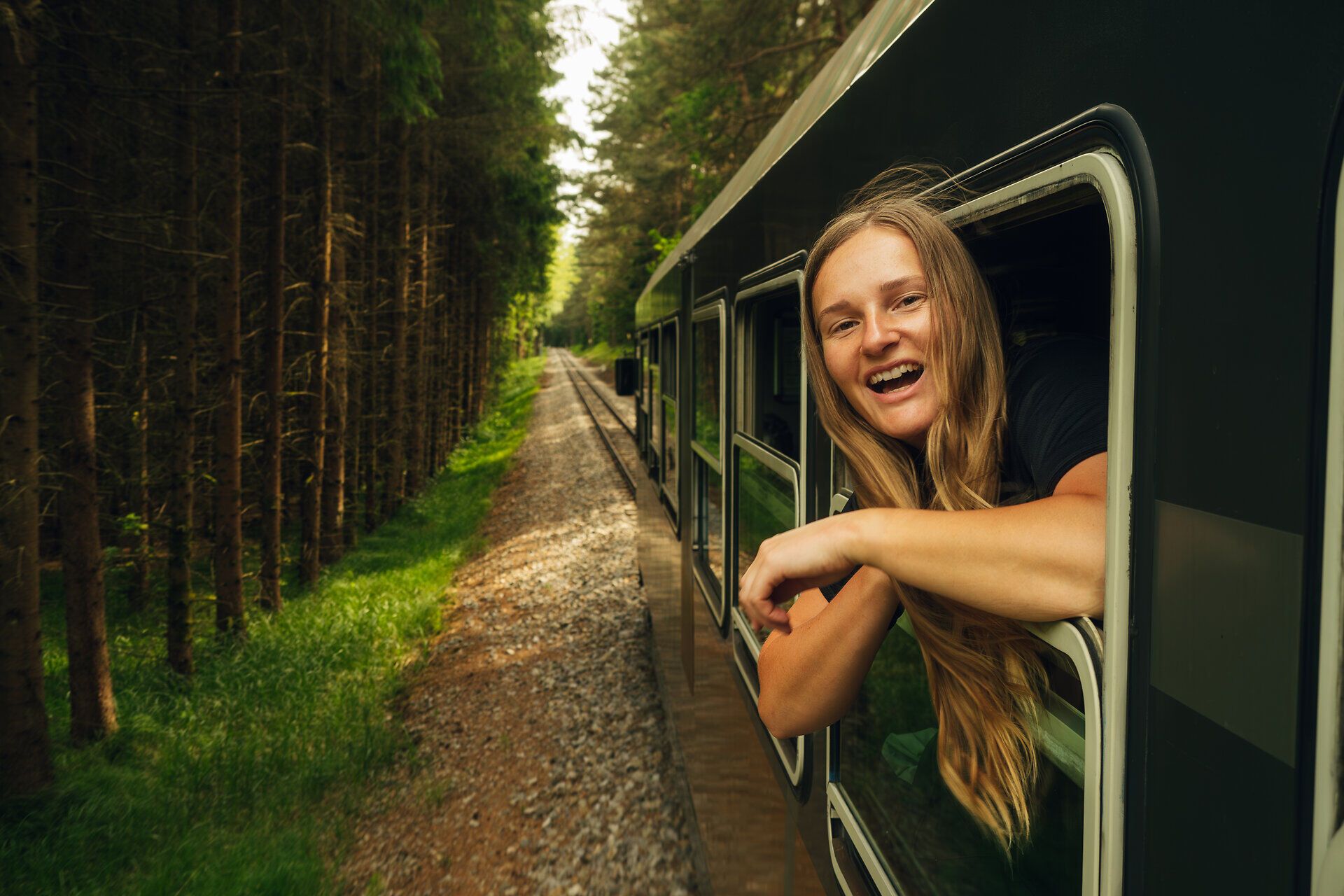 Eine lächelnde Frau lehnt sich aus dem Fenster eines fahrenden Zuges, der durch einen dichten Wald im Waldviertel fährt.