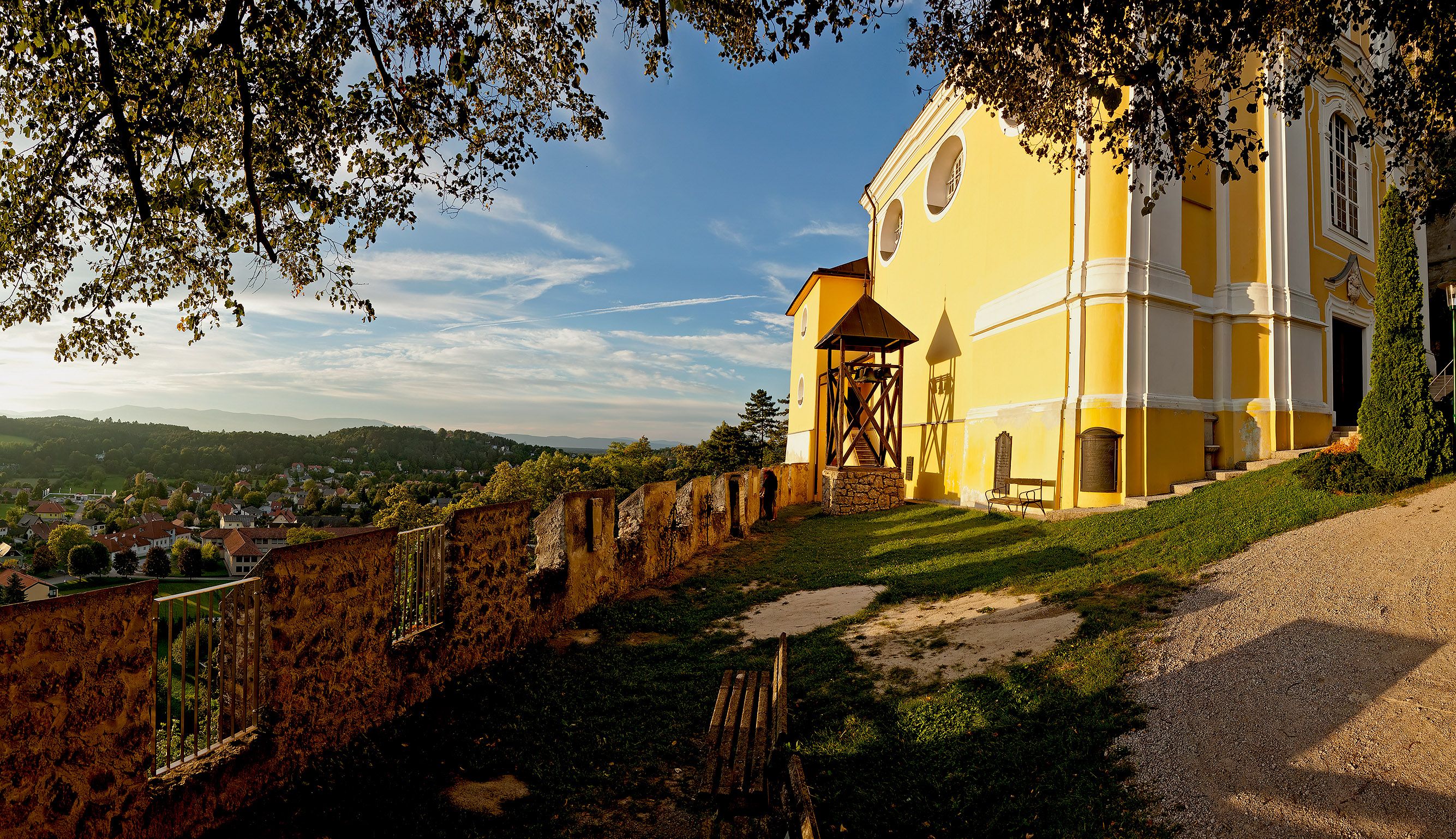 Gelbe Kirche auf einem Hügel mit Blick auf ein Dorf und bewaldete Hügel im Hintergrund.