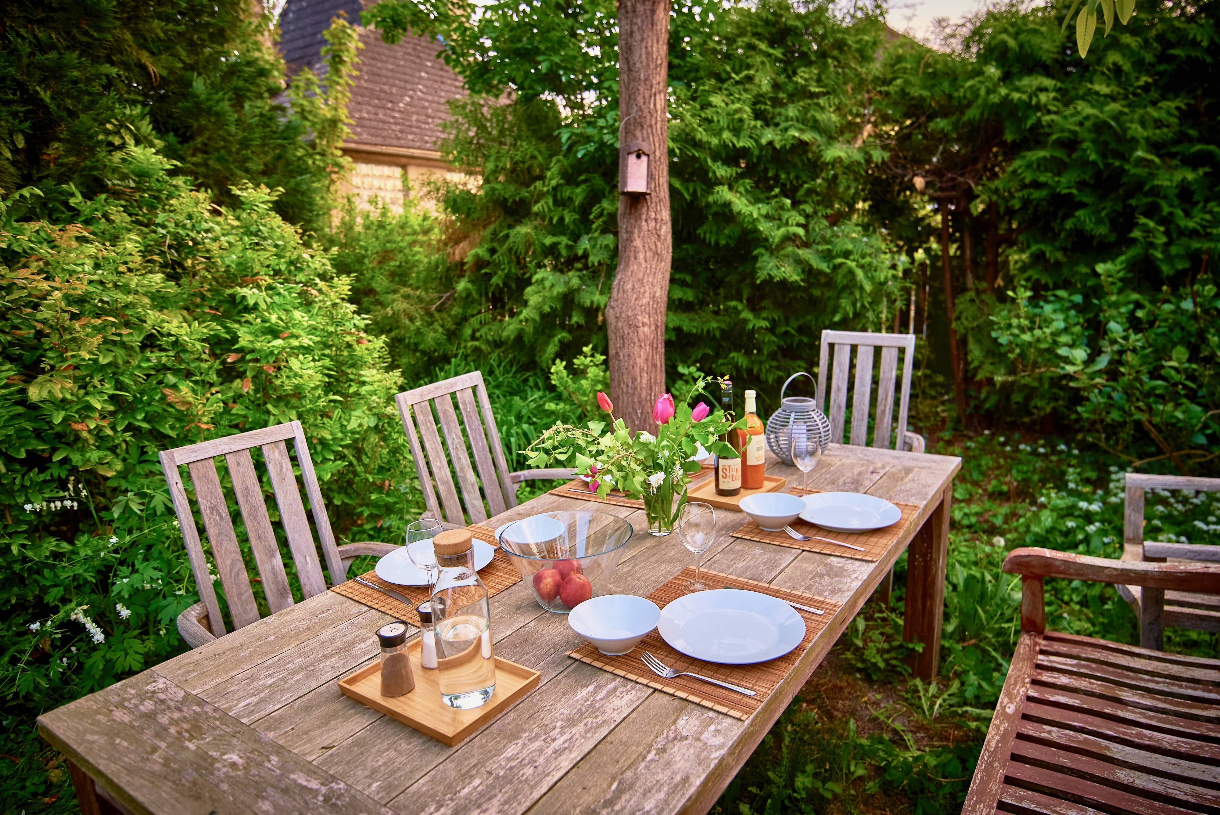 Ein gedeckter Holztisch im Garten mit Tellern, Gläsern, einer Vase mit Blumen und einer Flasche Wein.