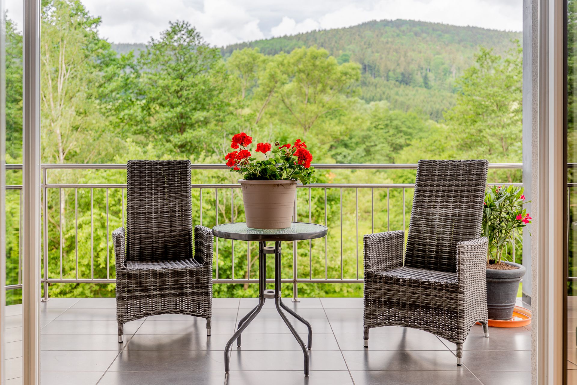 Balkon mit zwei Rattanstühlen, einem Tisch mit Blumentopf und Blick auf bewaldete Hügel.