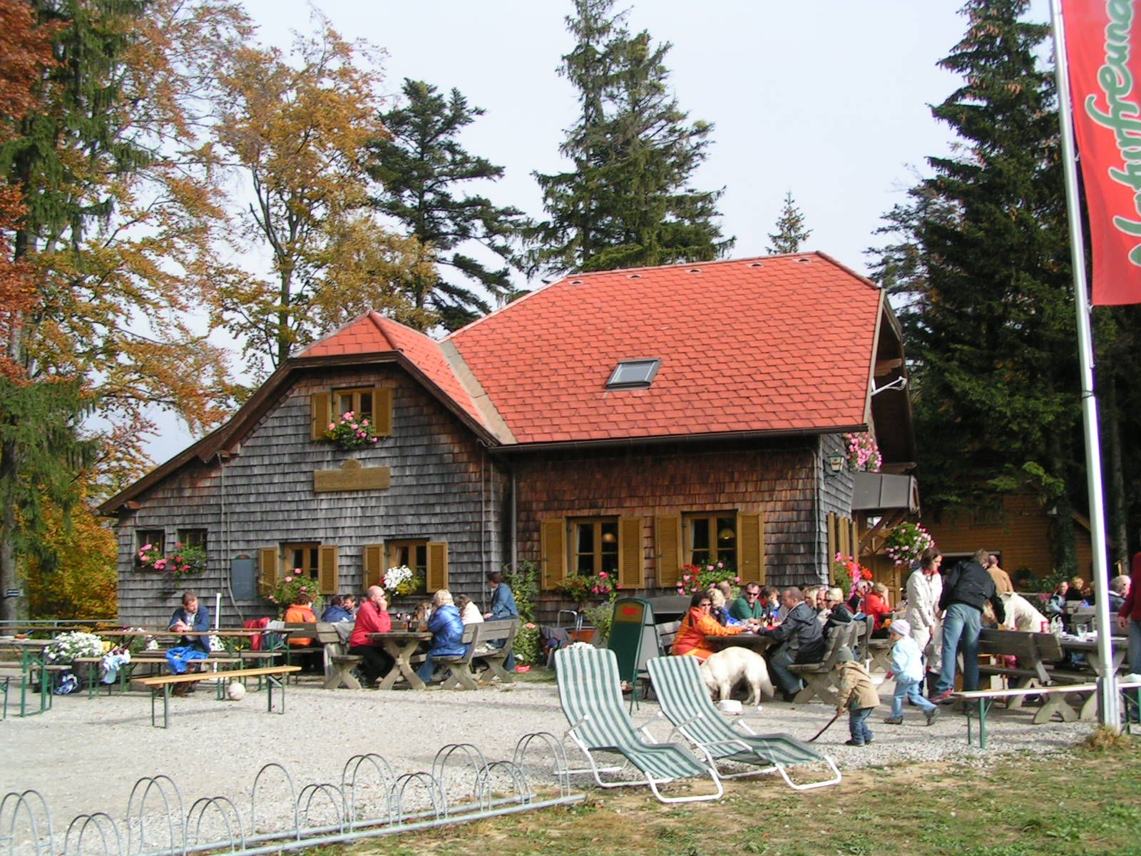 Eine Berghütte mit rotem Dach und Holzwänden, umgeben von Bäumen. Menschen sitzen draußen an Tischen, einige mit Hunden.