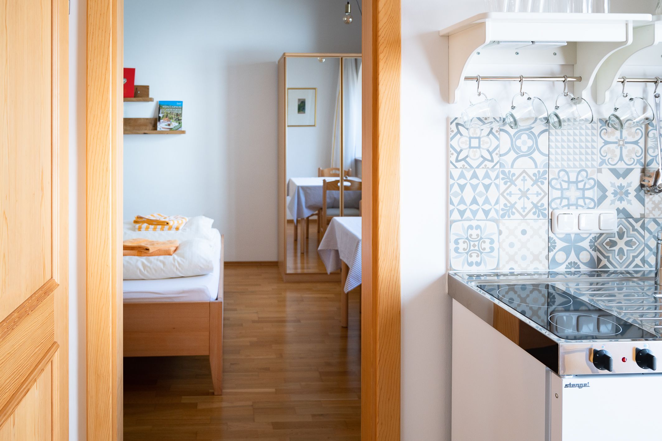 Innenansicht eines Zimmers mit Bett, Spiegel, Tisch und kleiner Küche mit gemusterten Fliesen.