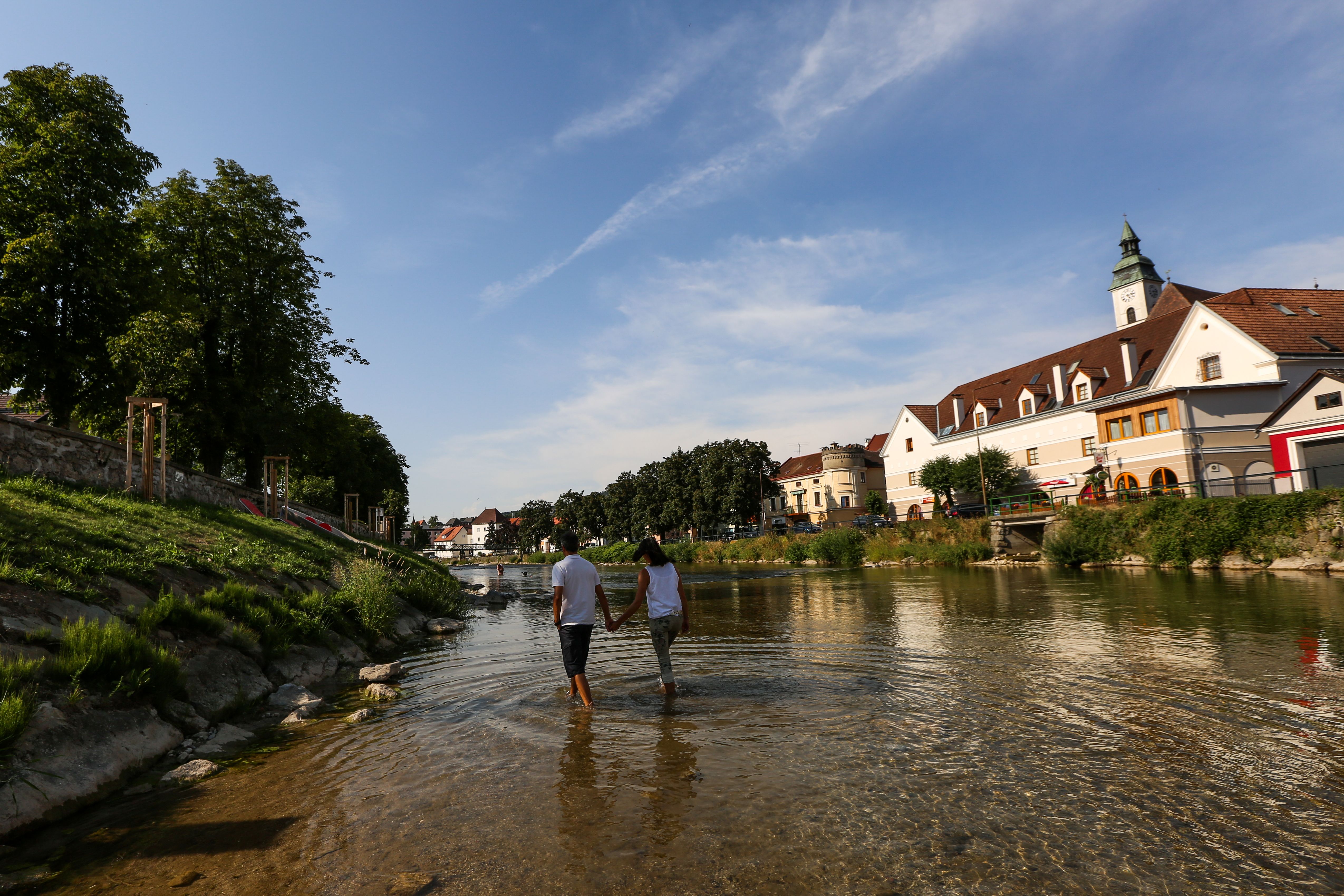 Ein Paar geht Hand in Hand durch einen flachen Fluss in einer Stadt mit historischen Gebäuden und Bäumen.