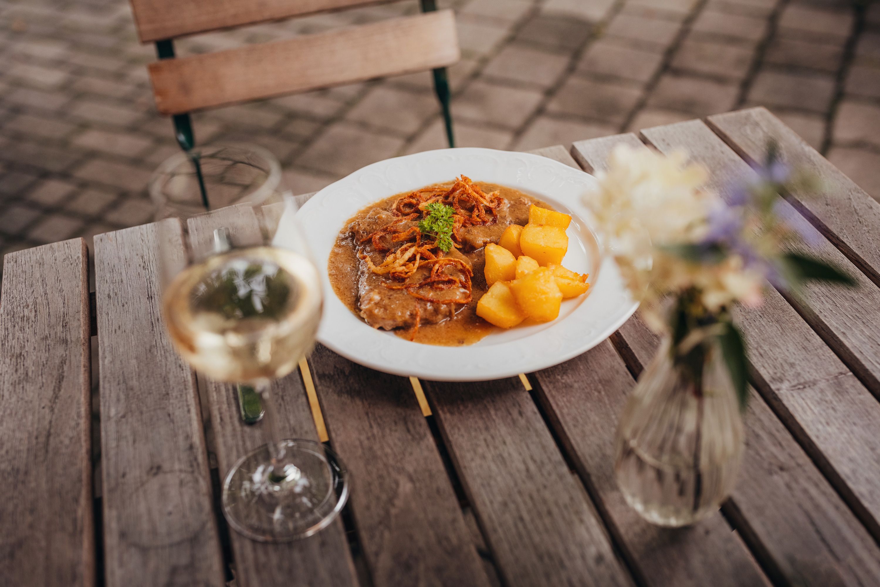 Ein Teller mit Zwiebelrostbraten und Braterdäpfeln auf einem Holztisch, daneben ein Glas Weißwein und eine Blumenvase.