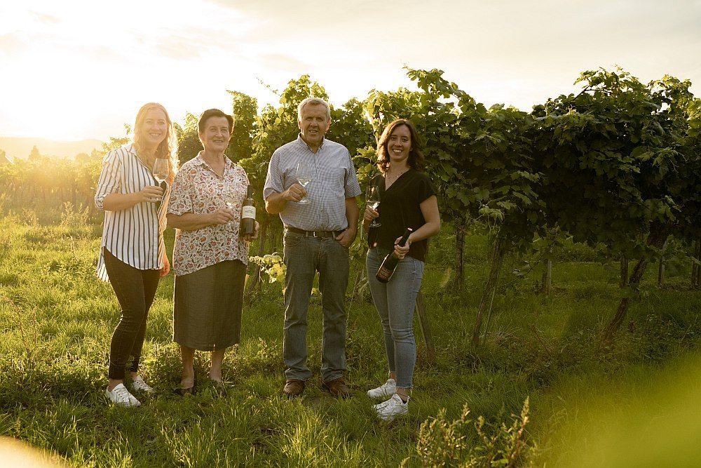 Vier Personen stehen in einem Weinberg und halten Weingläser und Flaschen in der Hand.