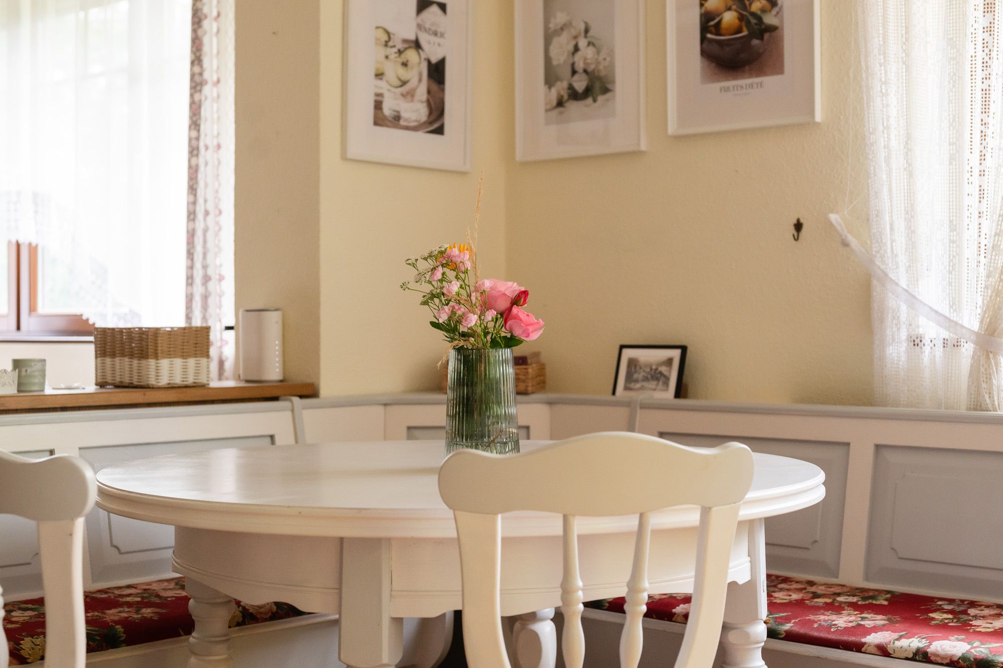 Essbereich in einem Ferienhaus mit rundem Tisch, Blumenstrauß in Vase und dekorativen Bildern an der Wand.