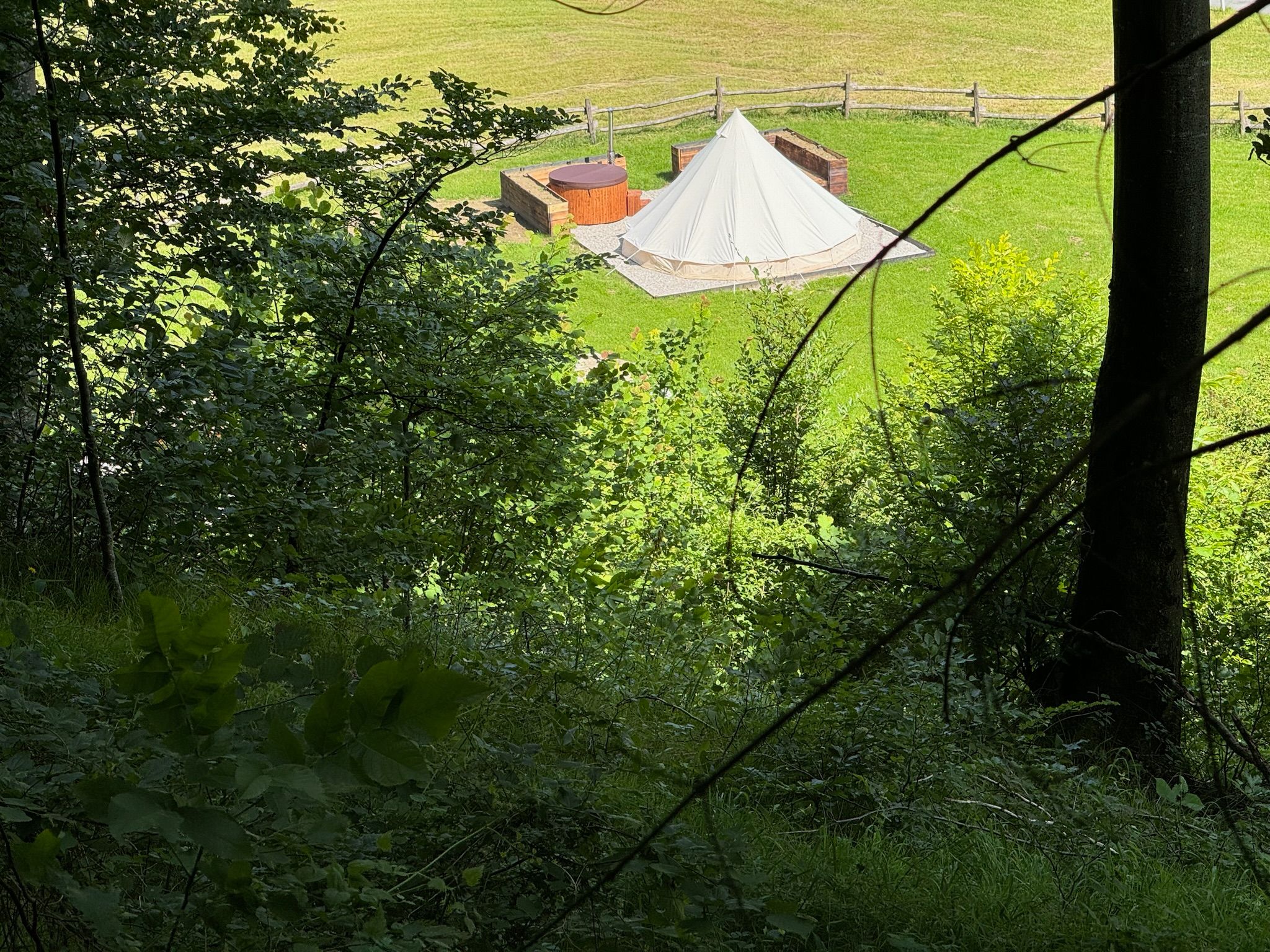 Blick auf ein Glamping-Zelt und einen Whirlpool auf einer Wiese, umgeben von Bäumen.