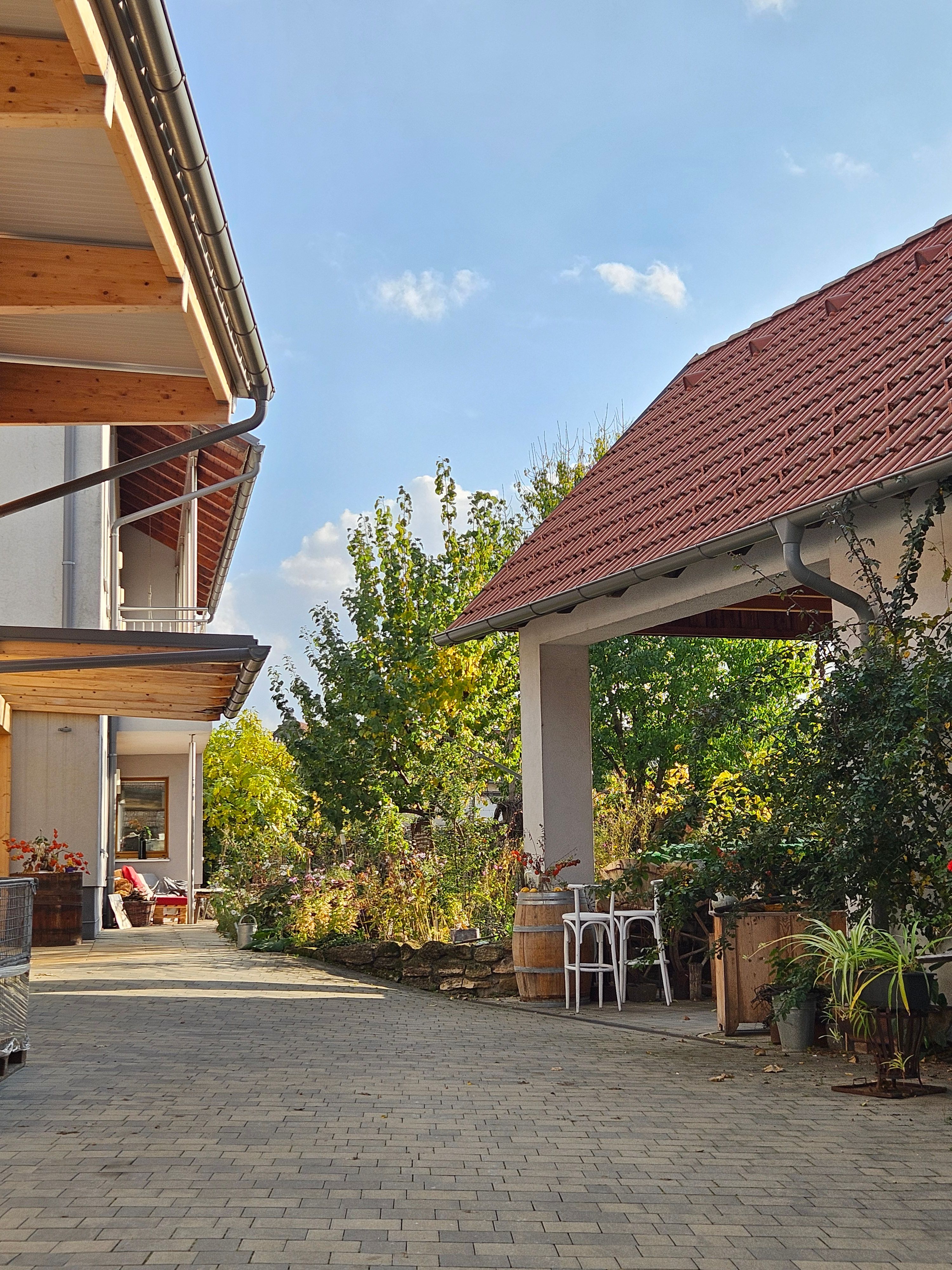 Ein gepflasterter Hof mit einem Gebäude und einem überdachten Bereich, umgeben von Pflanzen und Bäumen unter einem klaren blauen Himmel.