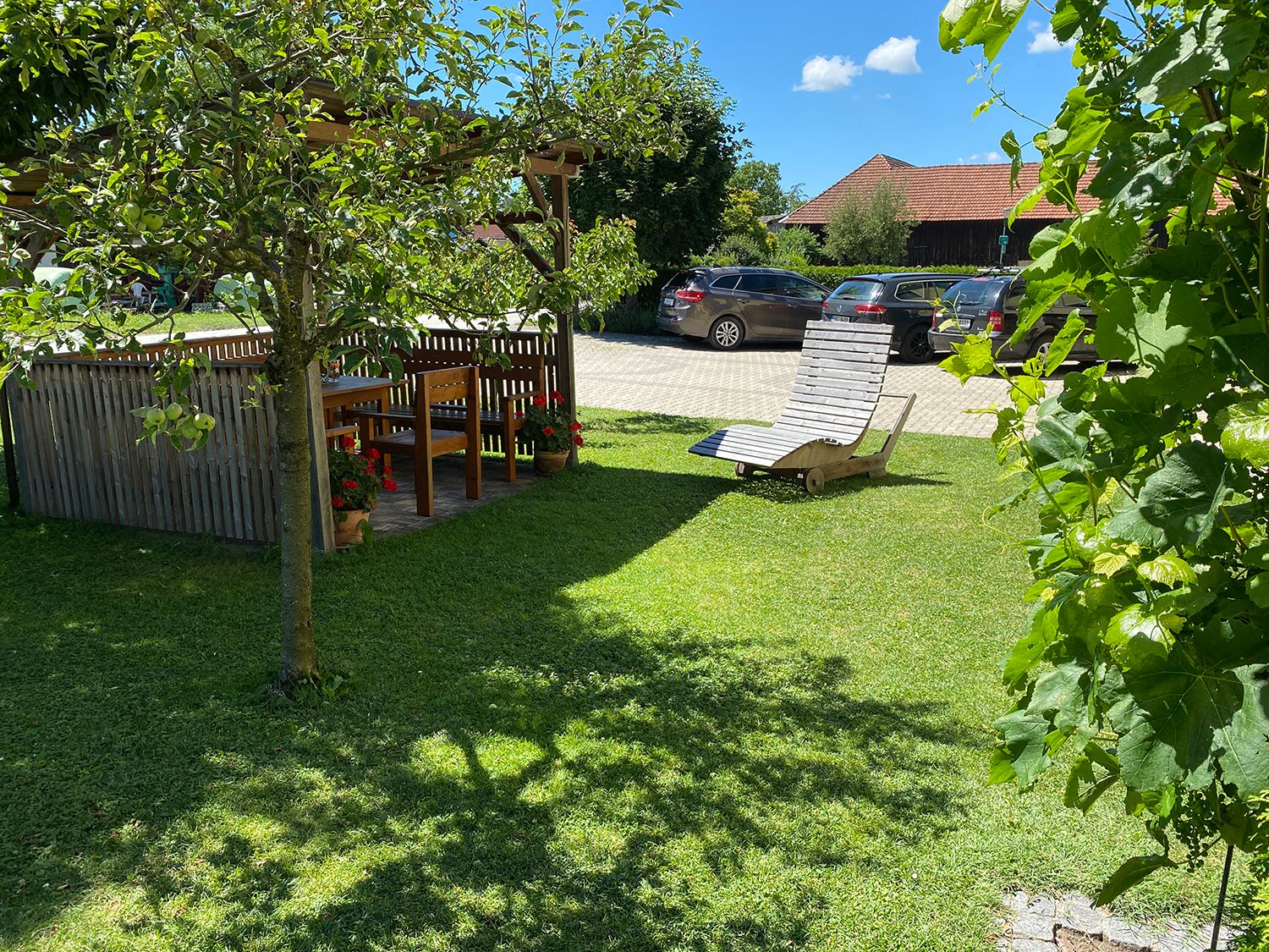 Garten mit Pergola, Holztisch und Stühlen, Liegestuhl auf Rasen, Autos im Hintergrund.