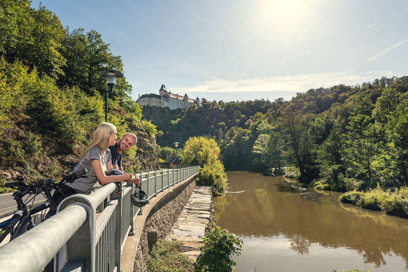 Zwei Personen lehnen an einem Geländer mit Blick auf einen Fluss und eine Burg im Hintergrund.