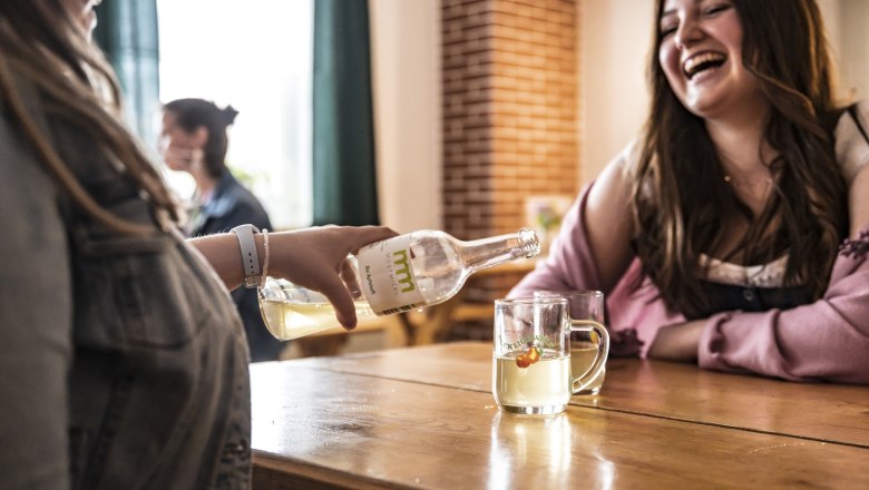 Zwei Frauen sitzen an einem Tisch, eine gie&szlig;t Most aus einer Flasche in ein Glas. Beide lachen.