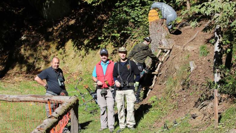 Drei M&auml;nner mit B&ouml;gen im Wald, ein Mann klettert eine Leiter hoch.
