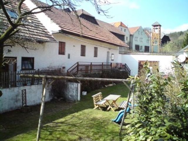 Ein ländlicher Hof mit weißen Gebäuden, Holztisch und Stühlen im Garten, Schaukel und Rutsche im Vordergrund.