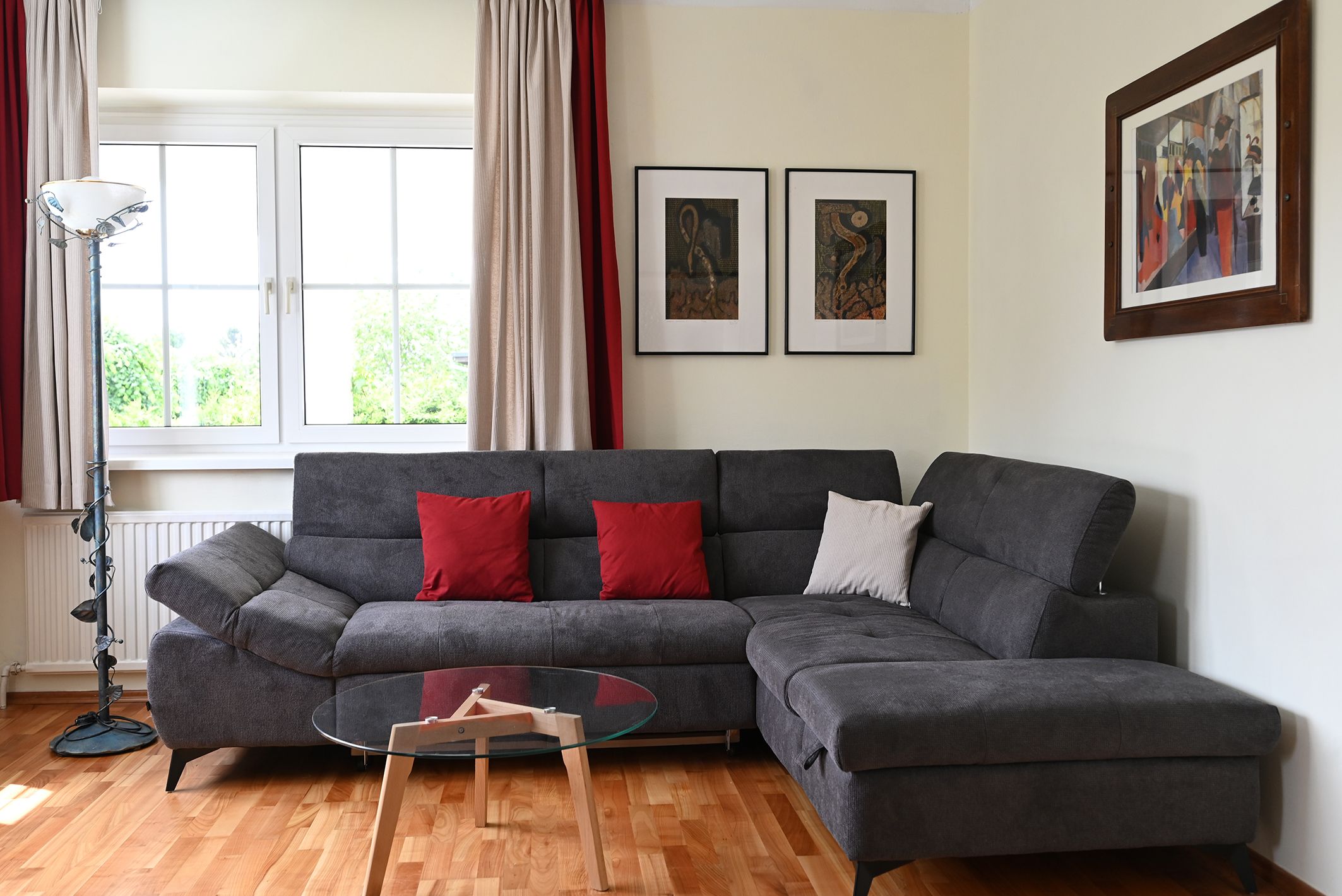 Modernes Wohnzimmer mit grauem Sofa, roten Kissen, Glastisch und Kunstwerken an der Wand.