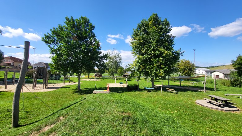 Ein Spielplatz mit Kletterger&uuml;st, Schaukeln und Picknicktisch auf einer gr&uuml;nen Wiese unter blauem Himmel.